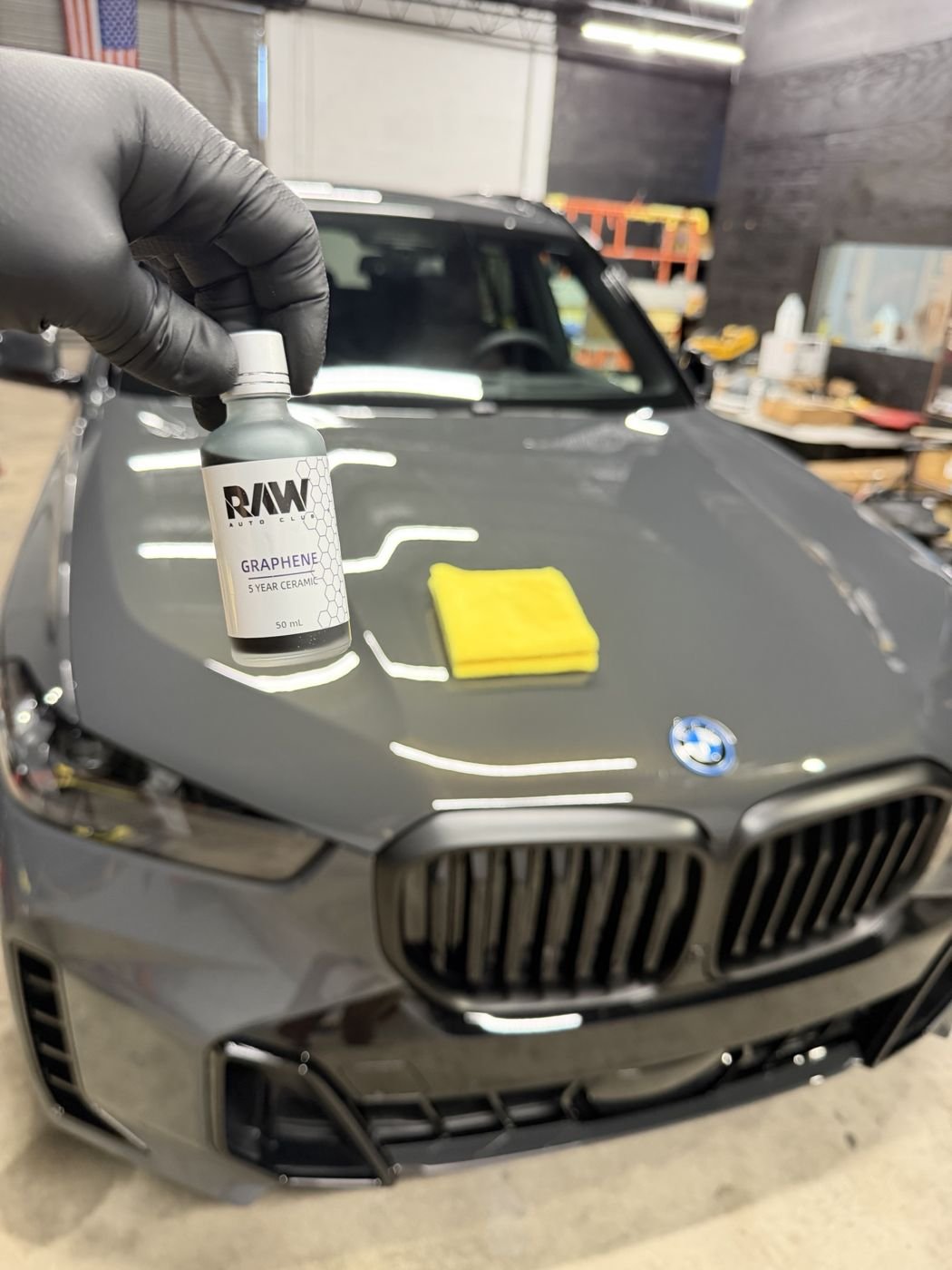 RAW Graphene ceramic coating bottle held over the freshly corrected hood of a gray BMW