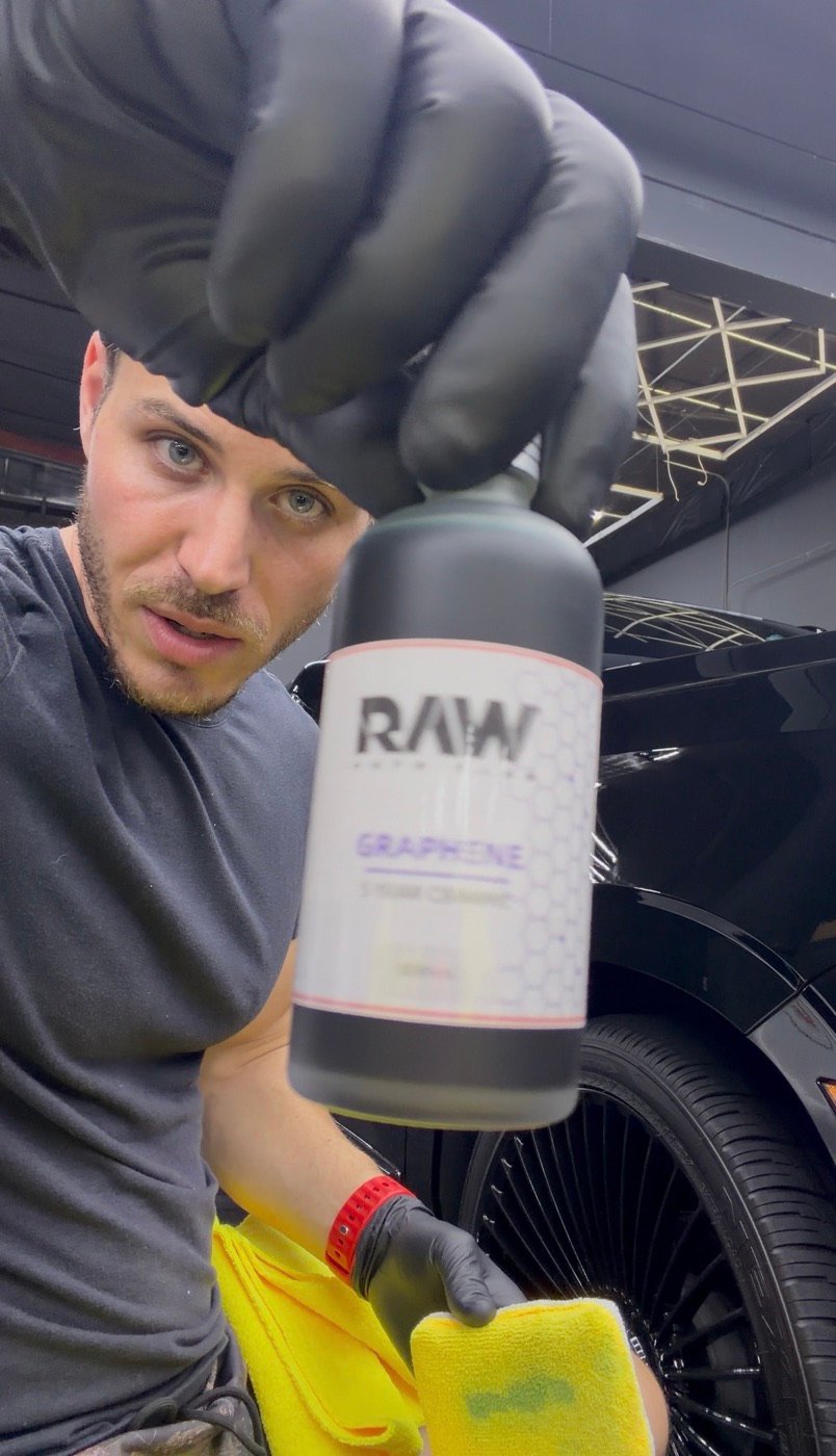Minty detailer holding a RAW graphene protection bottle beside a black vehicle